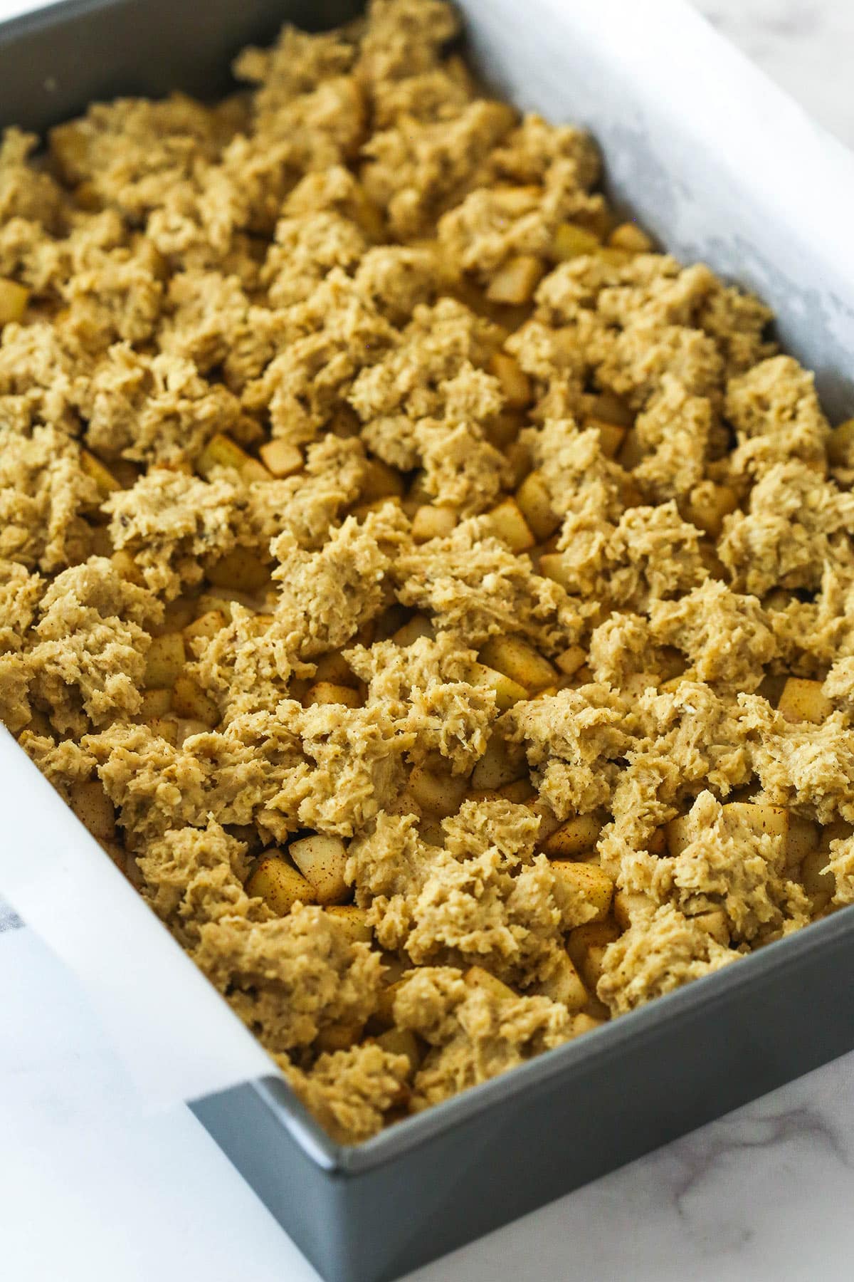 Oatmeal cookie dough crumbled on top of cinnamon apples in a baking pan.