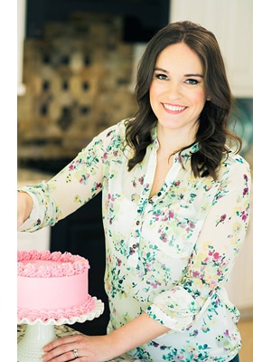 photo of lindsay in the kitchen standing with a pink cake on a cream colored cake stand
