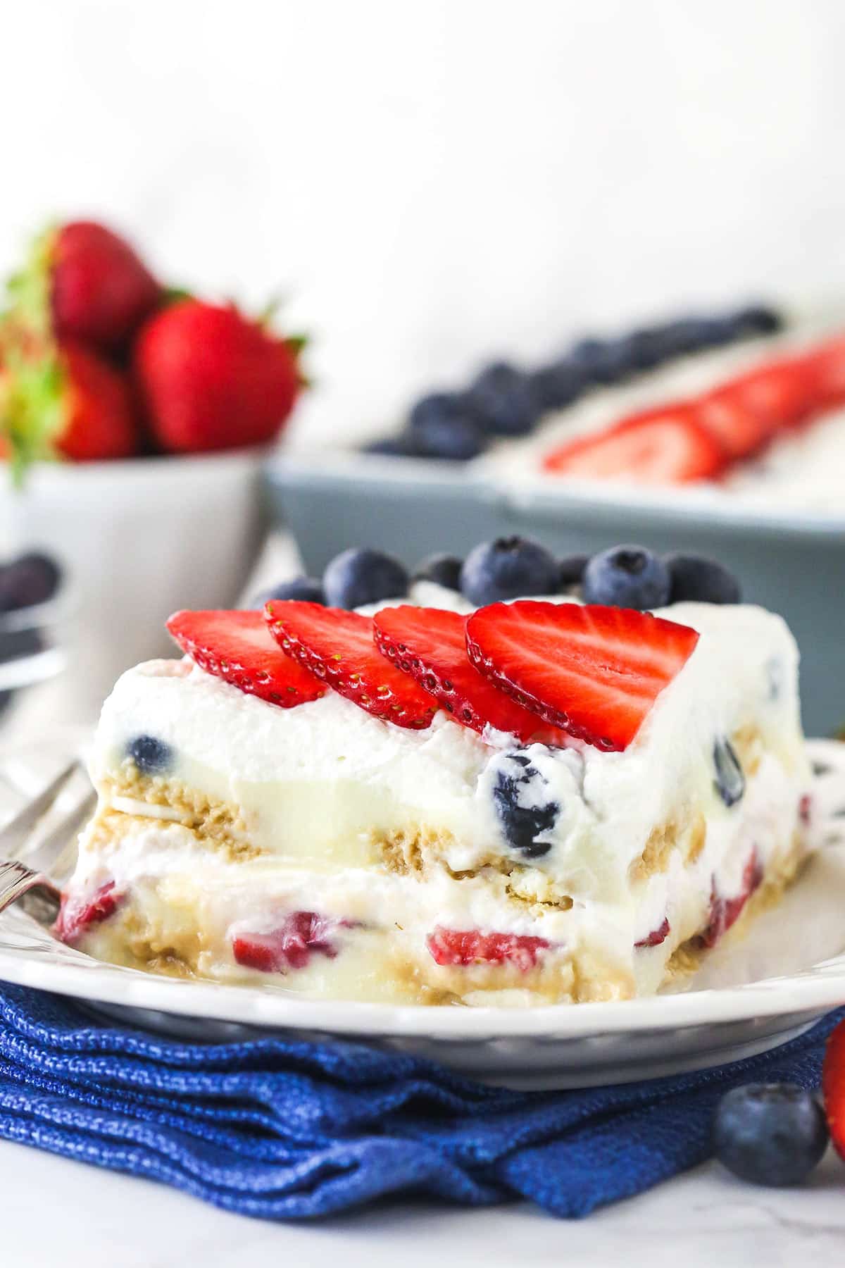 slice of berry icebox cake on white plate with blue napkin underneath and berries in the background