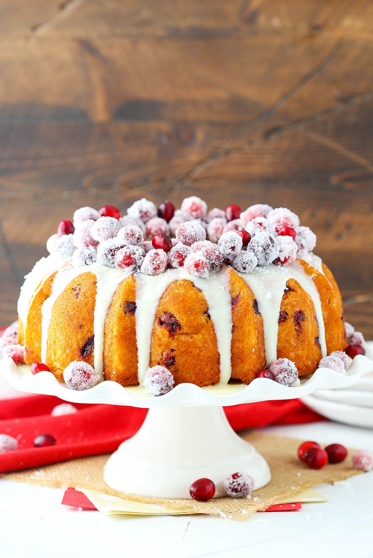 A completed cranberry white chocolate bundt cake on a dessert stand on top of a wicker placemat