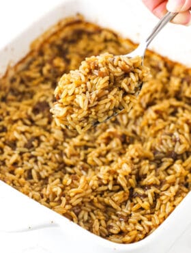 A metal spoon holding up a big bite of baked rice with a pan full of more rice beneath it