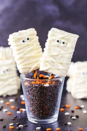 Two Mummy Rice Krispie Treats on orange and black straws in a glass filled with chocolate sprinkles.