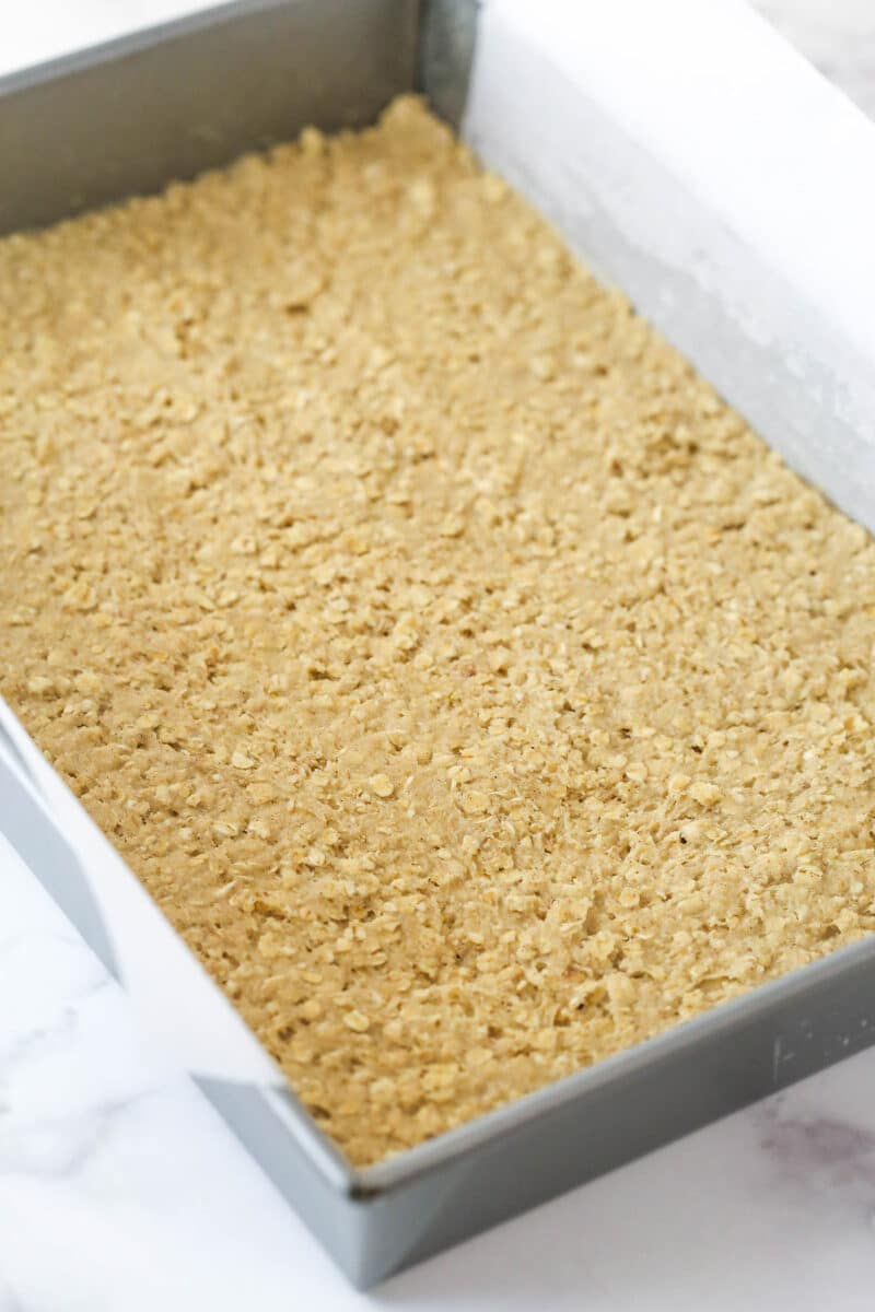 Pressing oatmeal cookie dough into a baking pan lined with parchment paper.