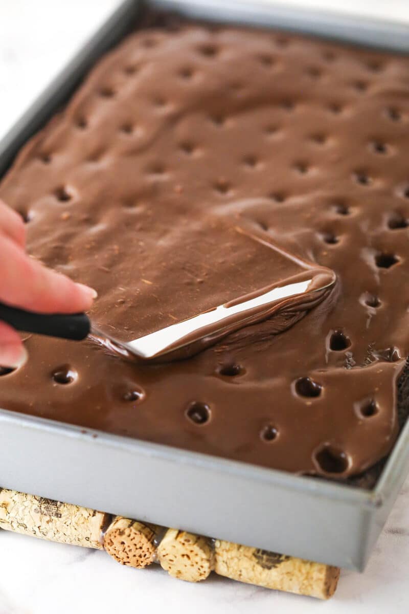 Spreading chocolate sauce over chocolate cake with holes poked in it.