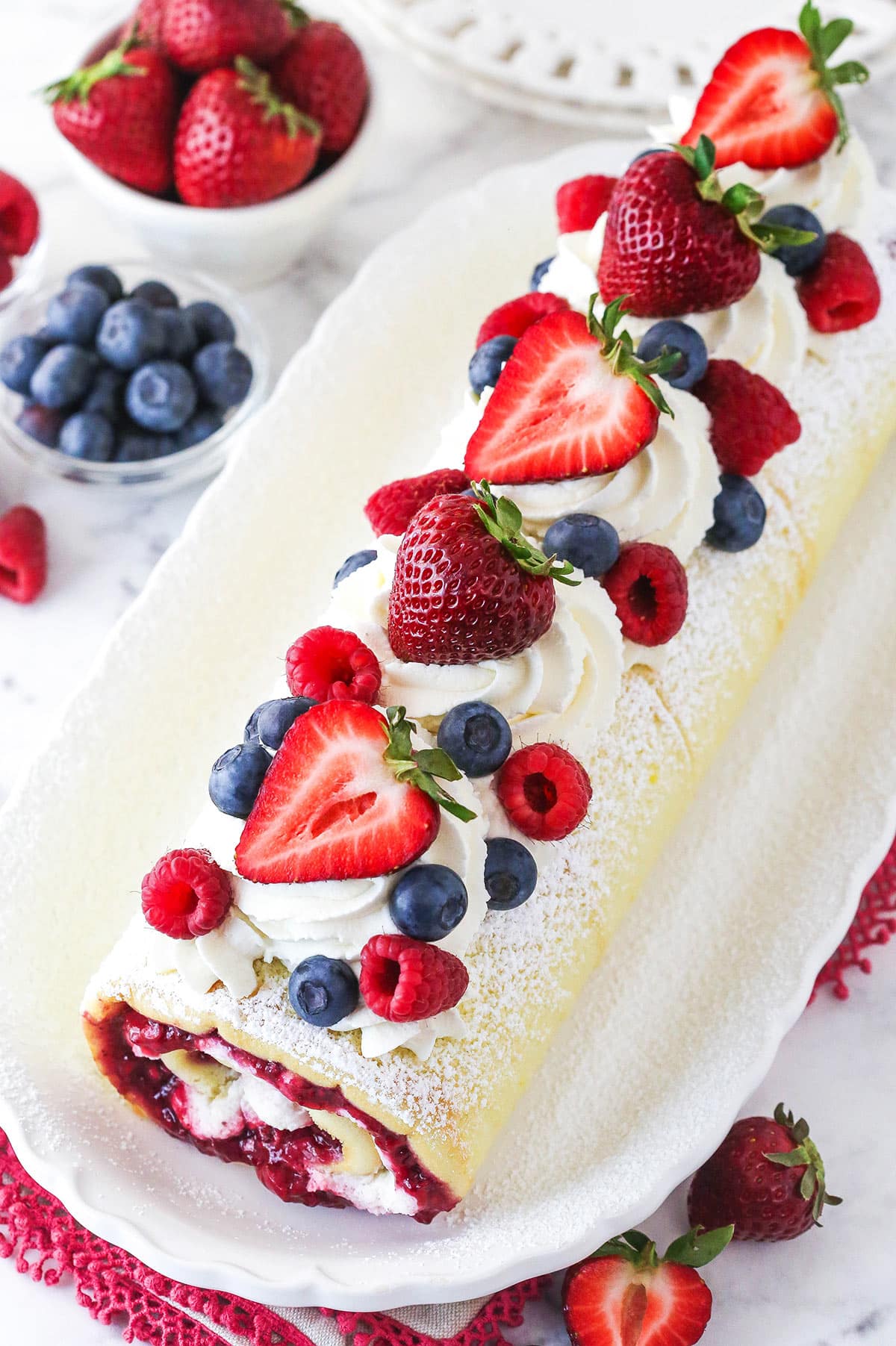 Berry Vanilla Cake Roll on white platter decorated with powdered sugar and berries, with berries in background