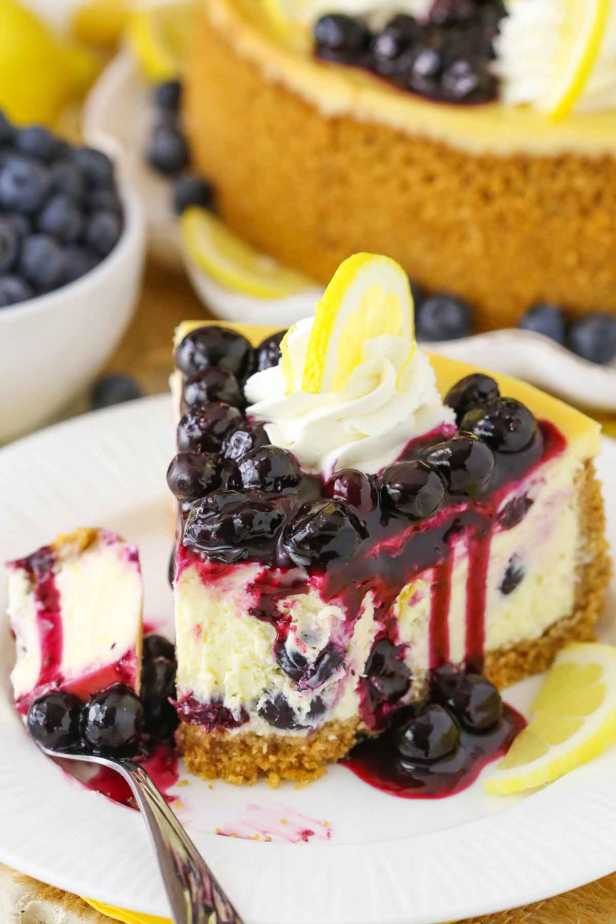 A slice of Lemon Blueberry Cheesecake with a bite removed on a white plate