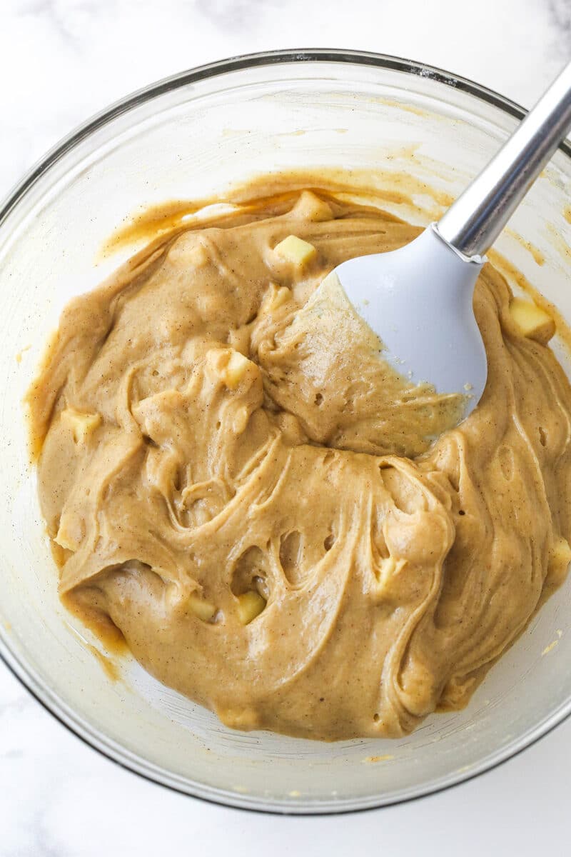 A glass mixing bowl filled with blondie batter that has apples mixed into it.