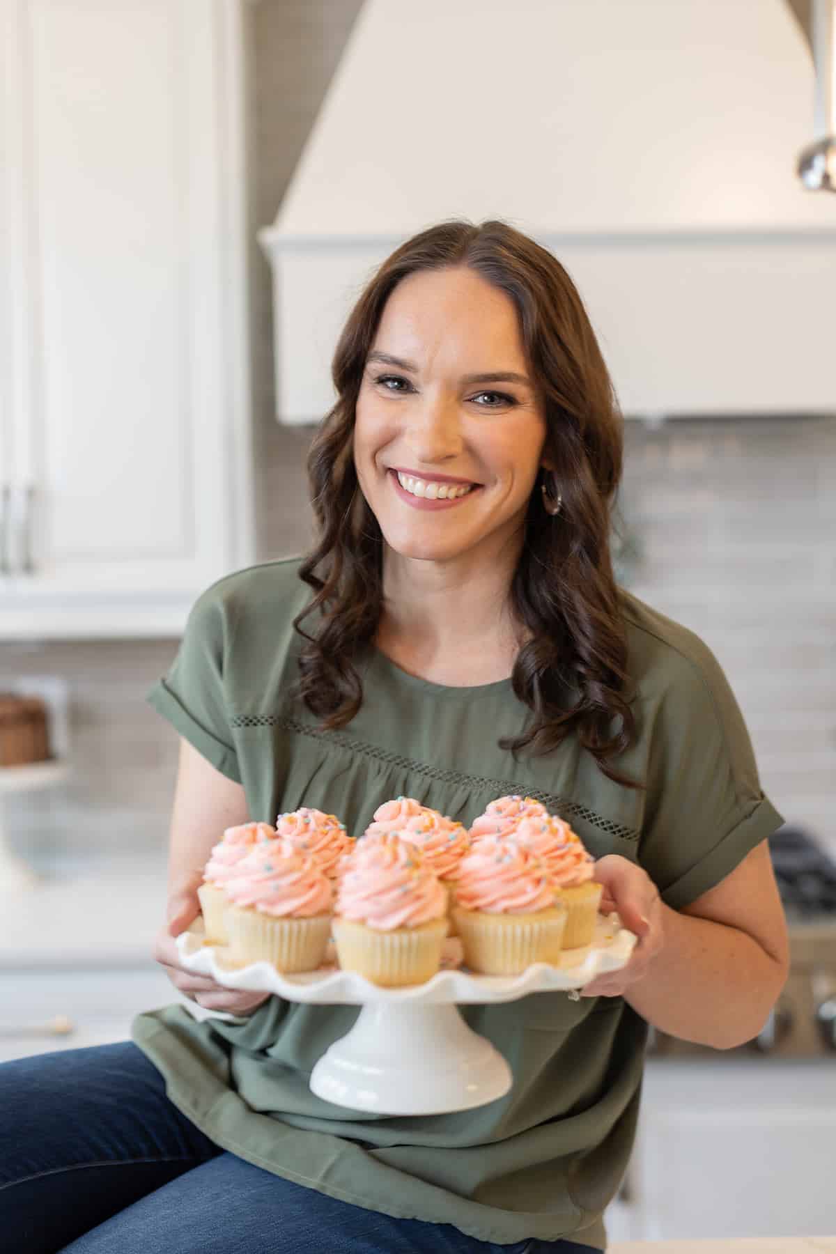 Lindsay sitting on a countertop holding a cake stand with cupcakes on it