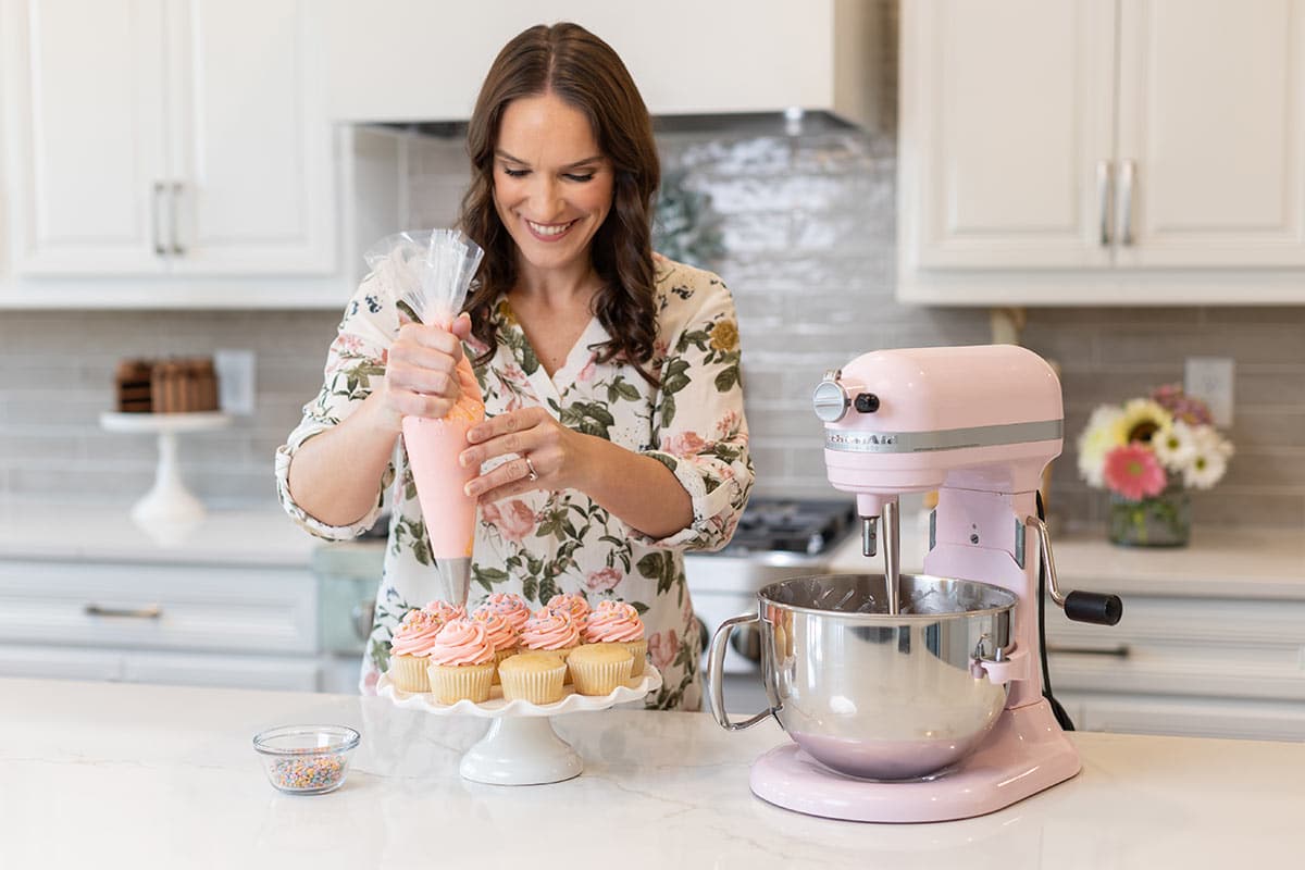 Lindsay piping pink frosting onto vanilla cupcakes with pink mixer next to her