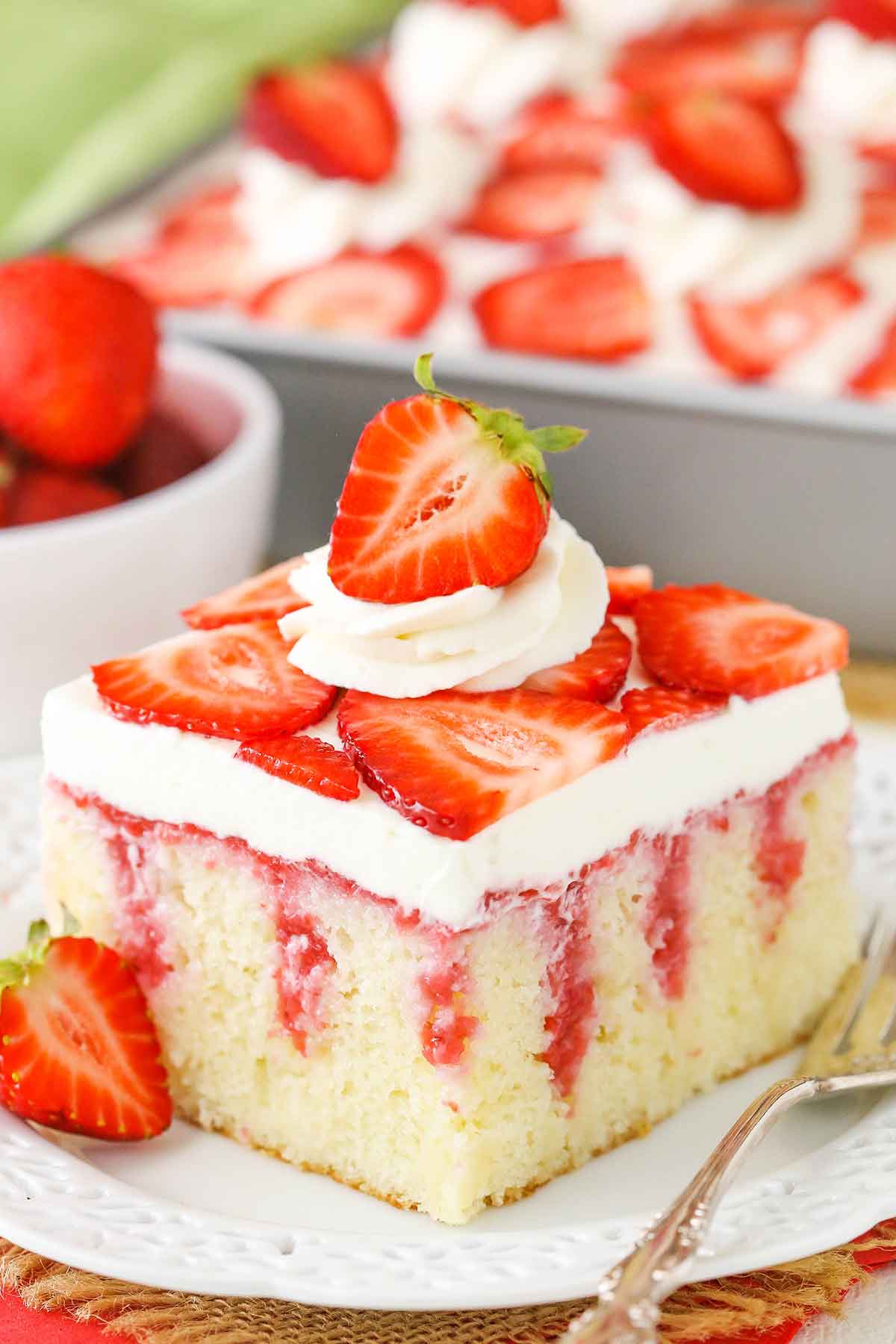 A square serving of Strawberry Poke Cake on a white plate next to a silver fork