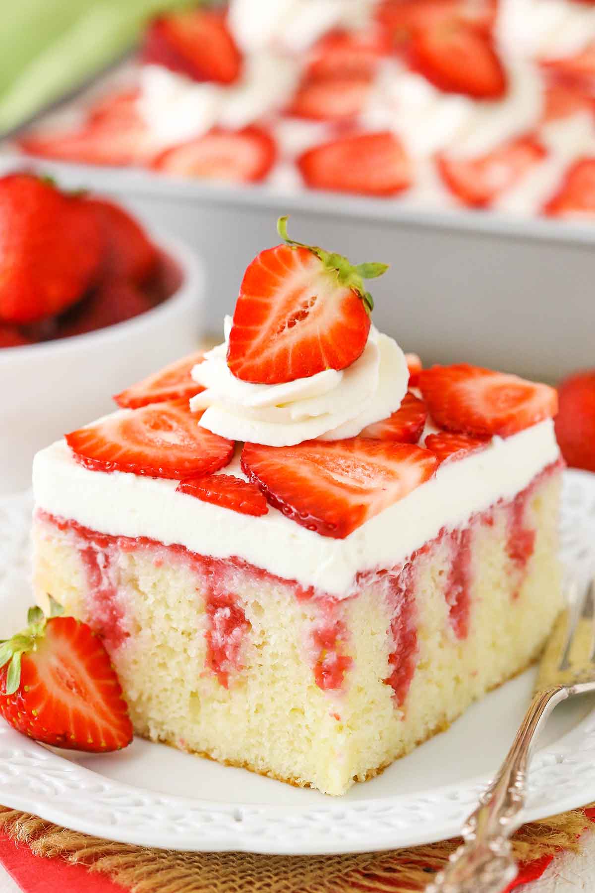 A square serving of Strawberry Poke Cake on a white plate next to a silver fork