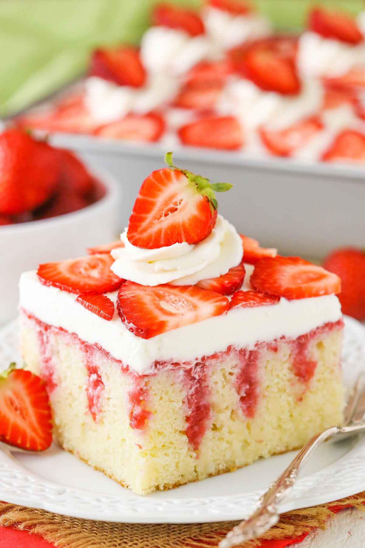 A square serving of Strawberry Poke Cake on a white plate next to a silver fork