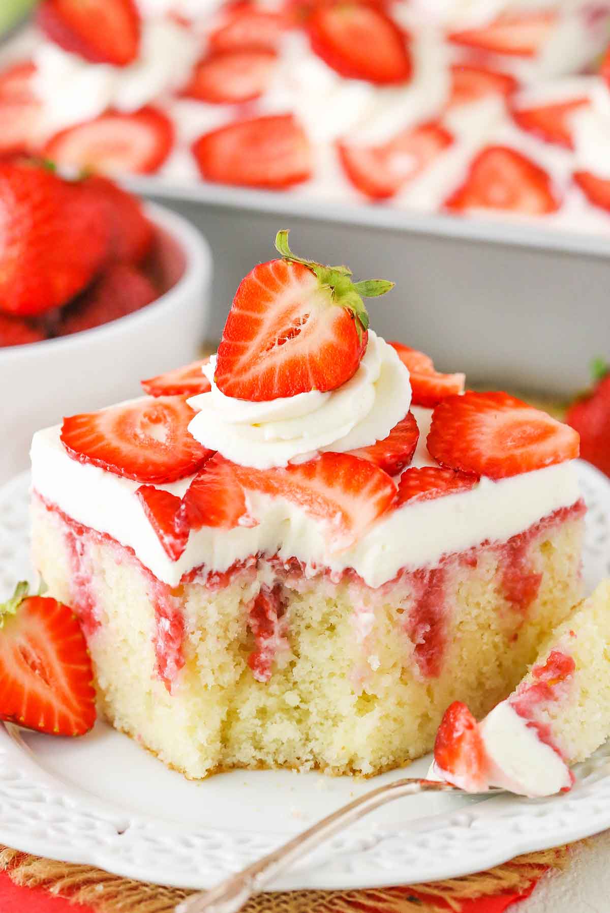 A square serving of Strawberry Poke Cake with a bite removed on a white plate next to a silver fork