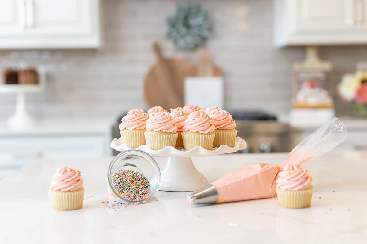 cupcakes, sprinkles and piping bag sitting on countertop with some cupcakes on a cake stand