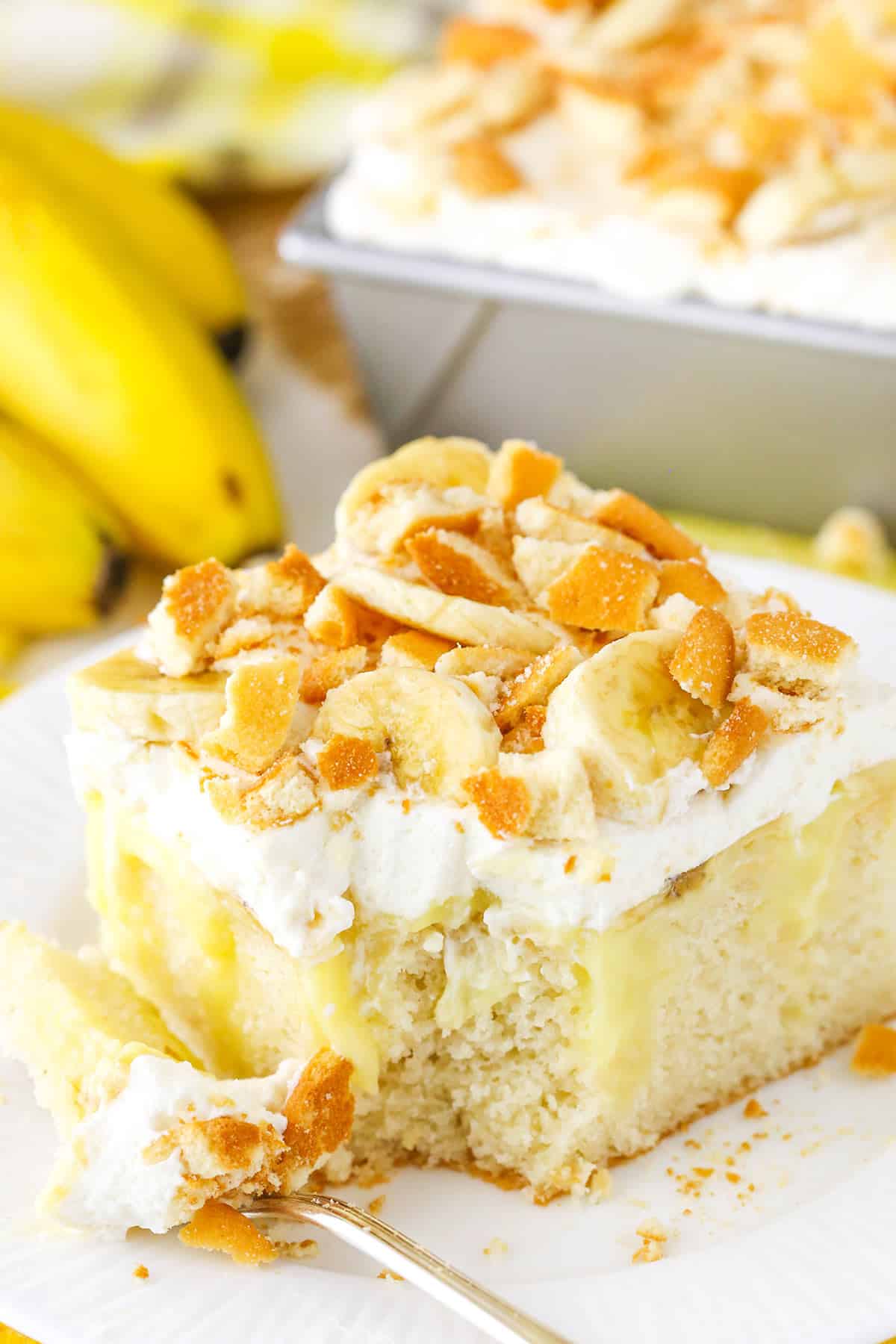 A square serving of Banana Poke Cake with a bite removed next to a fork on a white plate