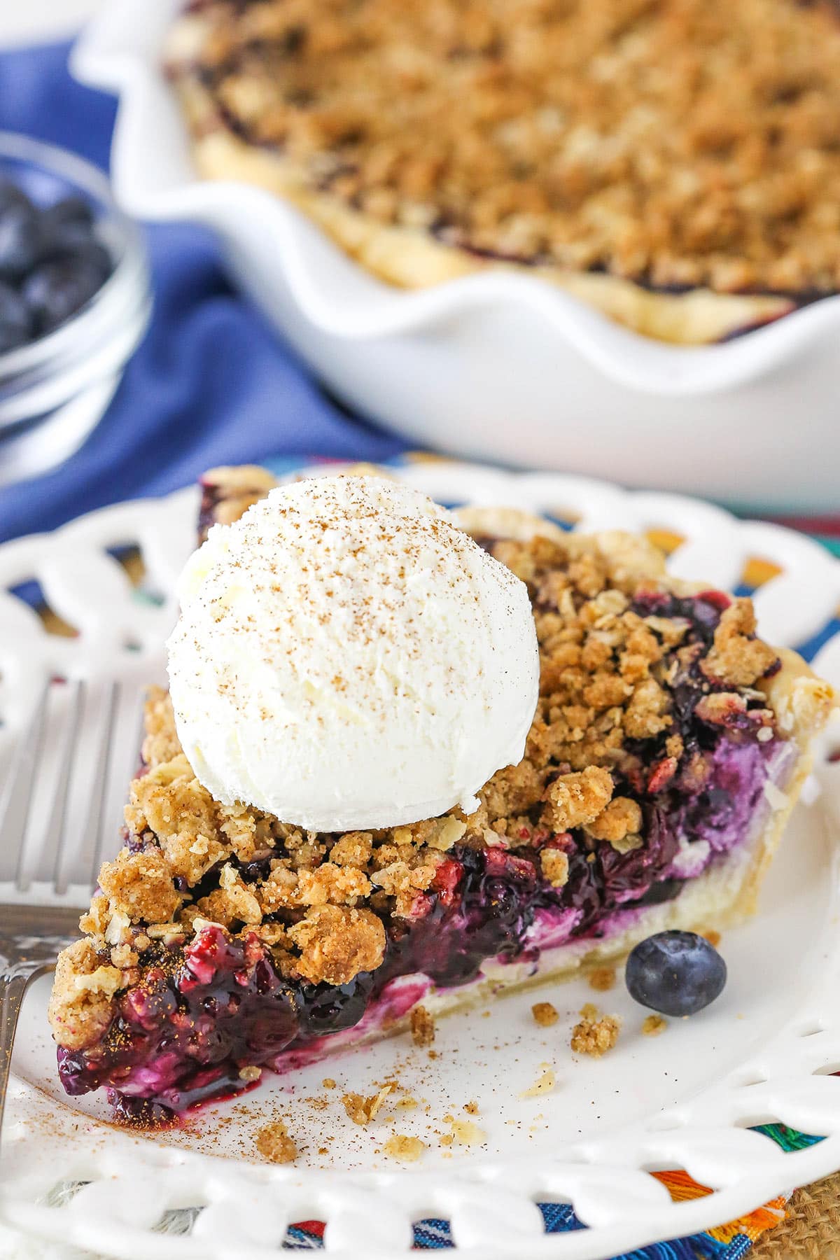 A slice of Blueberry Crumb Cheesecake Pie with a scoop of ice cream next to a fork on a white plate