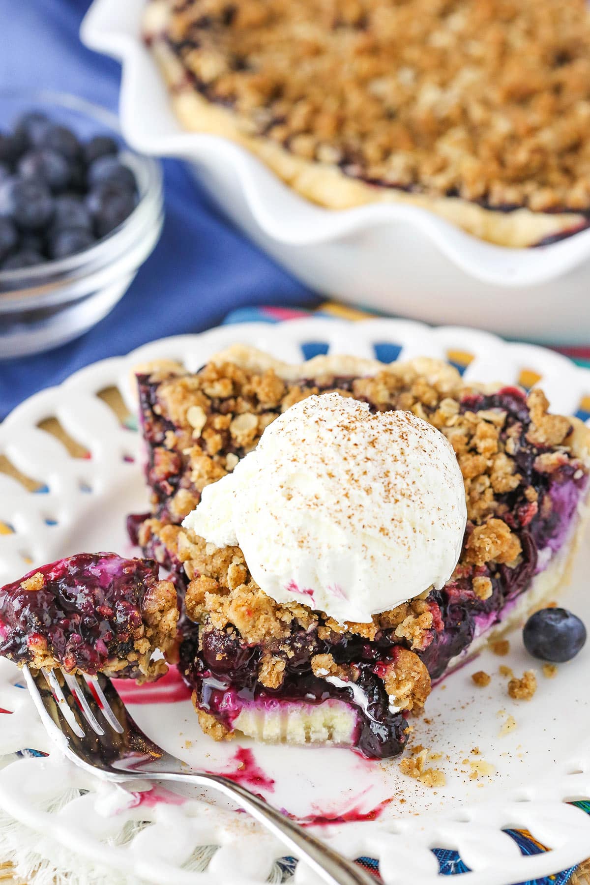 A slice of Blueberry Crumb Cheesecake Pie with a bite removed and a scoop of ice cream on top on a white plate