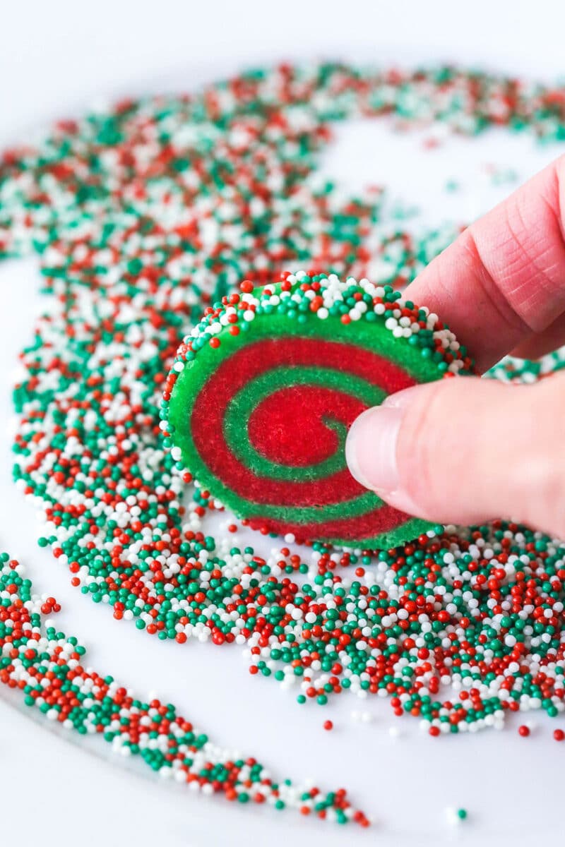 A hand rolling an unbaked pinwheel cookie in sprinkles